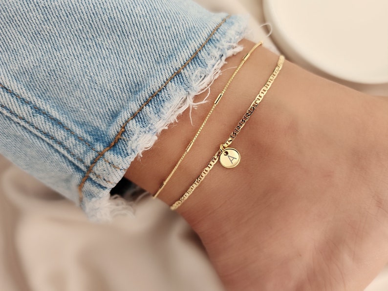Op de afbeelding: Gouden enkelband met een klein bedeltje met de letter 'A' erop. De enkelband is gelaagd met een dunnere ketting.