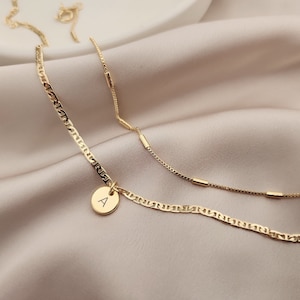 Op de afbeelding: Twee gouden kettingen, een met een delicate ketting en de andere met een dikkere, meer getextureerde ketting. De dikkere ketting heeft een klein rond hangertje met de letter 'A' erop.