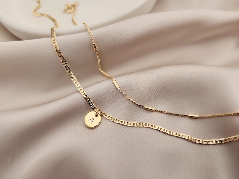 Op de afbeelding: Twee gouden kettingen, een met een delicate ketting en de andere met een dikkere, meer getextureerde ketting. De dikkere ketting heeft een klein rond hangertje met de letter 'A' erop.