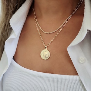May include: A gold chain necklace with a round pendant featuring a religious figure. The necklace is layered with a thinner gold chain.