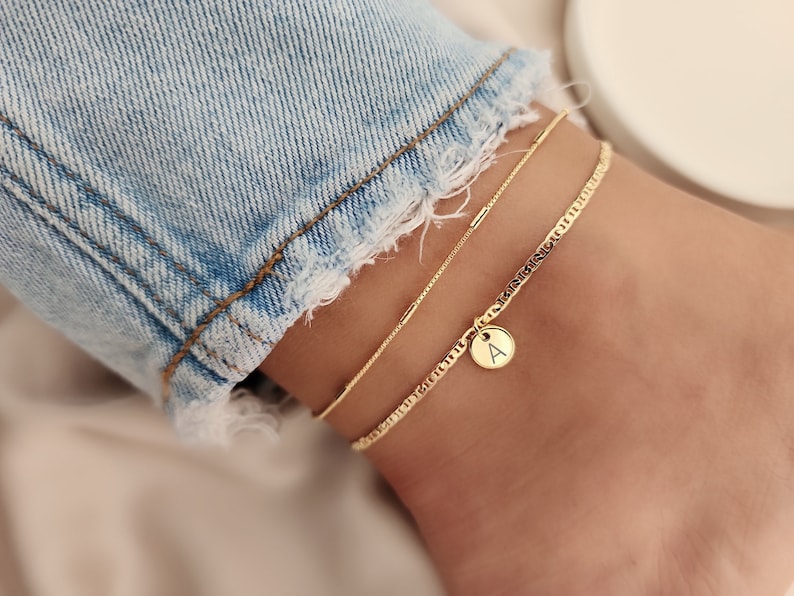 Op de afbeelding: Gouden enkelbandje met een delicate ketting en een bedel met de letter 'A'.