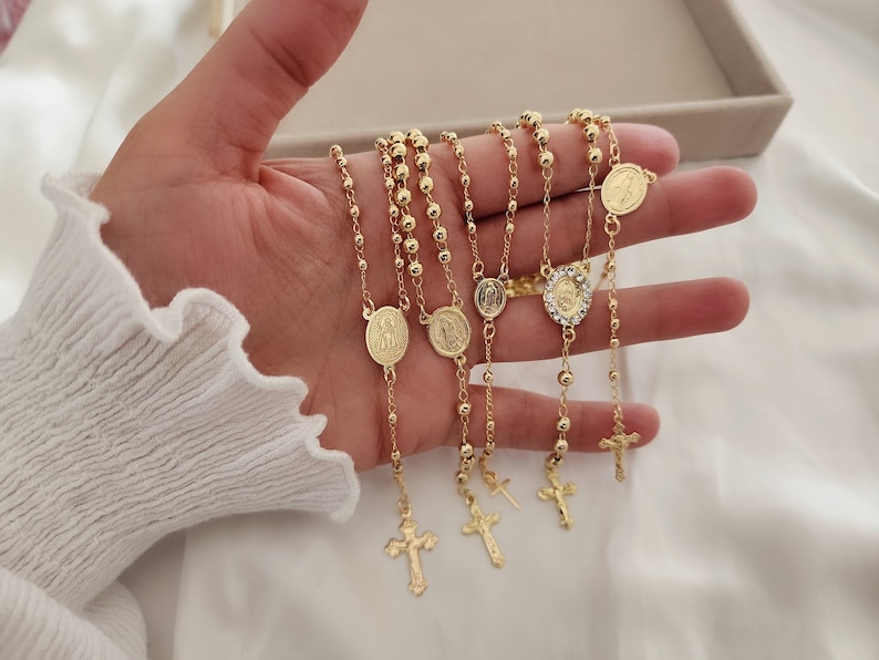 May include: Five gold chain necklaces with religious pendants. Each necklace has a different pendant, including a cross, a heart, and a round pendant with a face. The chains are made of small, round beads.