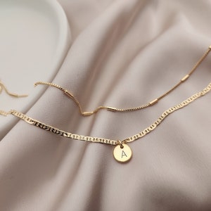 Op de afbeelding: Een gouden ketting met een klein gouden hanger met de letter 'A'. De ketting is gelaagd over een dunnere gouden ketting met kleine gouden staafjes.