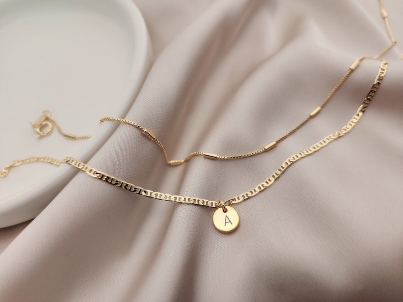 Op de afbeelding: Een gouden ketting met een klein gouden hanger met de letter 'A'. De ketting is gelaagd over een dunnere gouden ketting met kleine gouden staafjes.