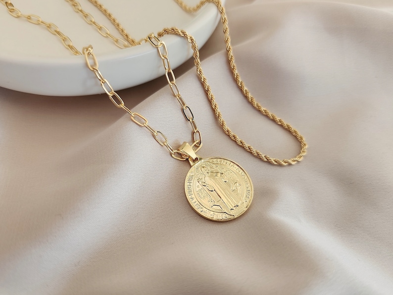 May include: A gold chain necklace with a round pendant featuring a religious image. The necklace is layered with a rope chain.