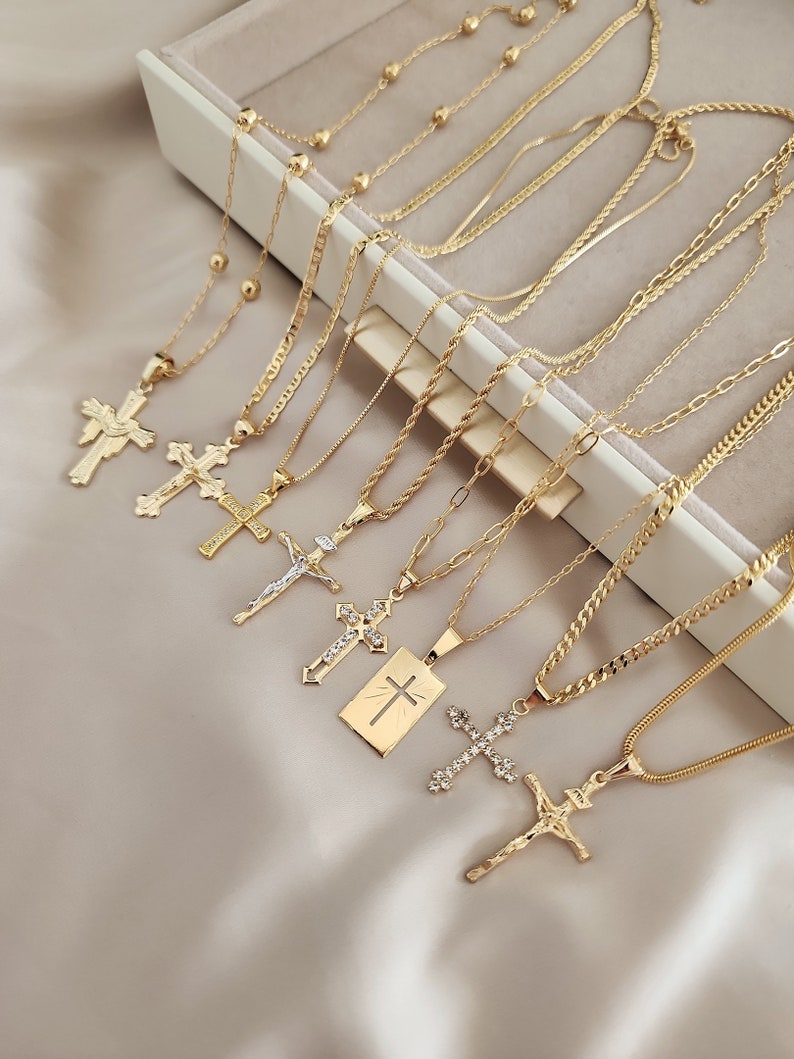 May include: A set of gold chain necklaces with various cross pendants. The chains are layered and feature different styles, including a beaded chain, a rope chain, and a curb chain. The cross pendants are all gold and feature different designs, including a simple cross, a cross with a figure of Jesus, and a cross with a sunburst design.