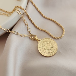 May include: A gold chain necklace with a gold pendant featuring a Saint Benedict medal. The medal has a cross and Latin text.