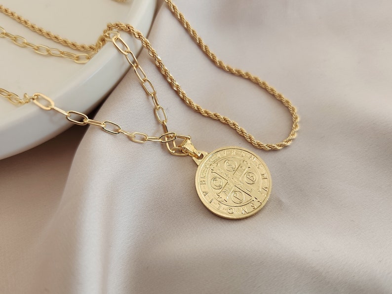 May include: A gold chain necklace with a gold pendant featuring a Saint Benedict medal. The medal has a cross and Latin text.