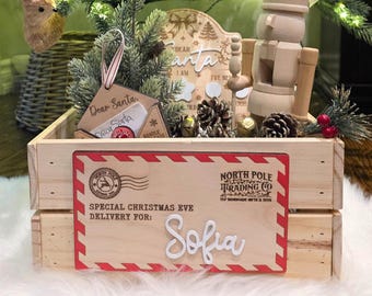 以下が含まれることがあります： 白い文字で「Sofia」と書かれた木製のクリスマスイブ配達ボックス。ボックスには赤と白のボーダーがあり、「North Pole Trading Co.」と「Special Christmas Eve Delivery For:」というテキストがあります。
