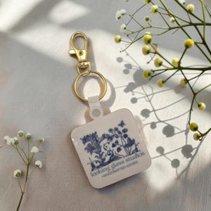 May include: A white square keyring with a blue floral pattern and the text "looking glass studios HANDCRAFTED GOODS". It features a gold-coloured clasp and ring.