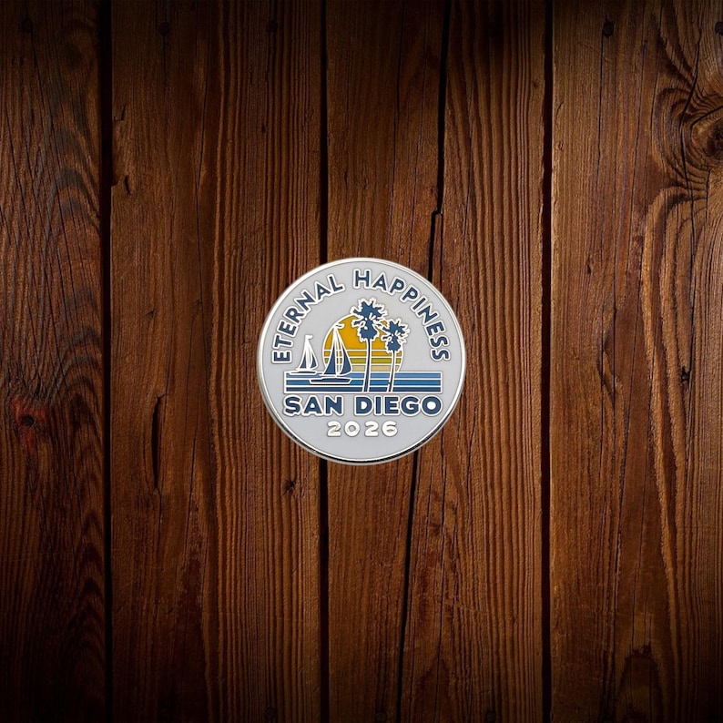 May include: A silver-colored round pin with the words "Eternal Happiness" and "San Diego 2026" encircling a design of a sailboat, sun, and palm trees. The pin is set against a dark brown wooden background.