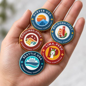 Puede incluir: Cinco pines circulares de esmalte con temas de convenciones internacionales. Los diseños incluyen Denver con una montaña, Bolonia con una torre y scooter, Coquimbo con uvas, Lima con una llama y Seattle con un ferry. Bordes dorados.