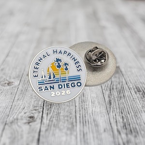 May include: A round, silver-colored pin with the words "Eternal Happiness" and "San Diego 2026" encircling a design of a sailboat, palm trees, and a sunset. The pin has a silver backing with a pin clasp.