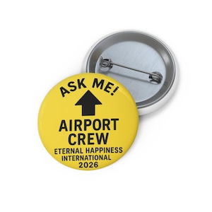 Może przedstawiać: Żółta przypinka z napisem "ASK ME!" nad czarną strzałką skierowaną w górę. Pod strzałką znajduje się napis "AIRPORT CREW" i "ETERNAL HAPPINESS INTERNATIONAL 2026". Przypinka ma metalowe zapięcie.