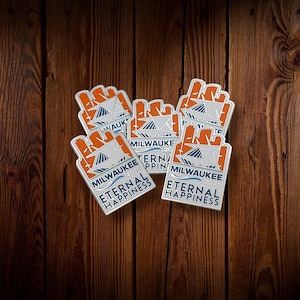 May include: A collection of rectangular enamel pins with a silver border. Each pin features an orange and white design with the text "MILWAUKEE 2026" and "ETERNAL HAPPINESS" in blue and white. The pins are arranged on a dark wood surface.