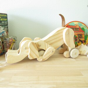 May include: A wooden toy dog with a pull string. The dog is made of natural wood and has a brown tail. The dog is lying on its side with its head turned to the left. The dog has four wheels and is ready to be pulled.