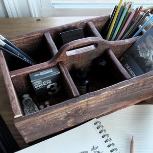 Könnte beinhalten: Ein Holzorganizer mit mehreren Fächern, gefüllt mit Künstlerbedarf wie Farbe, Pinseln, Bleistiften und einem Skizzenbuch. Der Organizer hat einen Griff und ist mit "Zeichnen" beschriftet.