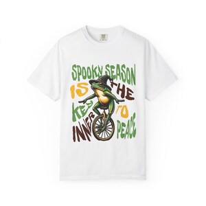 Puede incluir: Una camiseta blanca de manga corta con el gráfico de una rana verde con sombrero de bruja negro montada en un monociclo. El texto dice: SPOOKY SEASON IS THE KEY TO INNER PEACE, con letras en verde, amarillo y marrón oscuro alrededor.