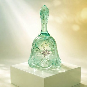 May include: A vintage-style, light green glass bell with a floral design. The bell features a long handle and a delicate, dangling purple flower. The bell is displayed on a white square pedestal.