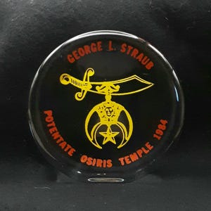 May include: A black, round emblem with the text "GEORGE L. STRAUB" in orange. A yellow sword and emblem are in the center, with "POTENTATE OSIRIS TEMPLE 1984" in orange around the edge.