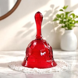 May include: A faceted, ruby-red glass bell with a long handle. The bell sits on a white, lace doily. A white vase with greenery is in the background. The bell's design features geometric shapes.