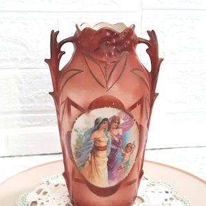 May include: A vintage ceramic vase with a pink and gold design. The vase features a painted scene of three women and a cherub. The vase has two handles and a scalloped rim.