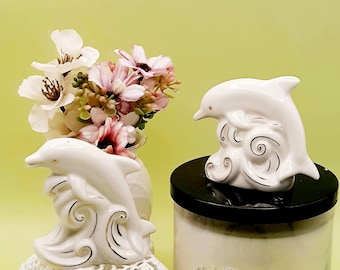 May include: White ceramic dolphin figurines, one atop a vase with flowers and the other on a black-topped container. The dolphins are detailed with gold accents and sit on stylized waves. The vase holds pink and white flowers.