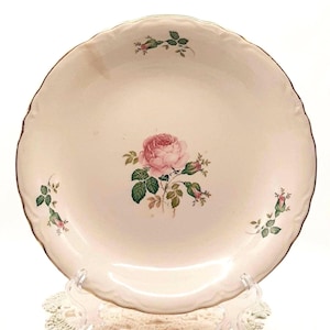 May include: A cream-colored, scalloped-edge plate with a pink rose and green leaves in the center. Additional rose buds and leaves are scattered around the plate. The rim has a gold trim. The plate is displayed on a clear stand.