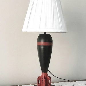 May include: A table lamp featuring a white pleated fabric lampshade atop a base shaped like a black and red bomb. The base includes a circular black stand with a red accent ring, a tapered black body with a red band, and red tail fins.