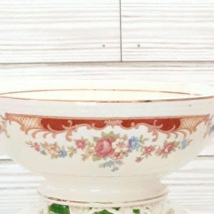 May include: A white porcelain bowl with a floral design and gold trim. The bowl has two handles and sits on a pedestal.