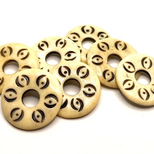 May include: Seven off-white bone beads with a carved design of eyes and crescent moons. The beads have a hole in the centre for stringing.