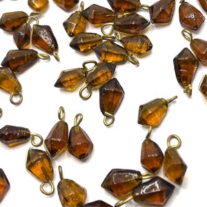 May include: A collection of amber glass teardrop-shaped beads with gold-toned metal loops. The beads are scattered on a white surface.