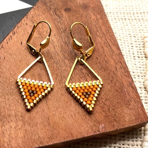 May include: A pair of gold-toned leverback earrings featuring a triangular pendant made of small, woven beads in shades of orange, yellow, and brown. The geometric design consists of a gold metal frame holding the beaded triangle at the base.