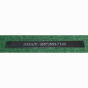May include: A black metal plate with the text "232A/V (657)333-7188" in a light color. The plate has two holes on each end. The plate is on a green surface.
