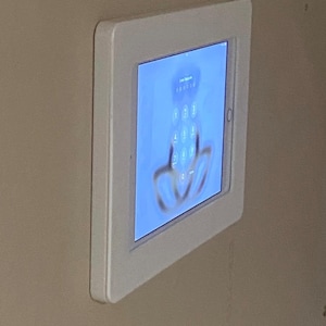 May include: A white, square tablet frame mounted on a beige wall. The tablet screen displays a blue interface with a keypad and a blurred fingerprint graphic. The frame has a clean, modern design.