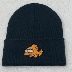 May include: Black knit beanie featuring an embroidered orange fish design. The fish has three eyes and the word "GCO" in white. The beanie is a classic, cuffed style, suitable for cold weather.
