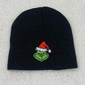 May include: Black knit beanie featuring an embroidered Grinch face wearing a Santa hat. The Grinch's face is green with yellow eyes, and the Santa hat is red with white trim. A winter accessory.