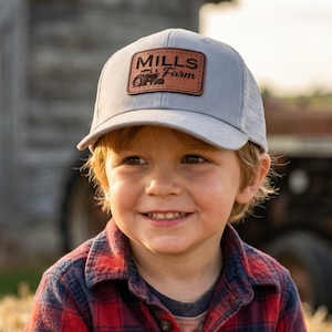 May include: A young person wearing a gray trucker hat with a brown leather patch that reads "Mills Farm". The person is also wearing a red and black plaid shirt. A tractor and hay bales are in the background.