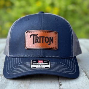 May include: A navy blue baseball cap with a grey mesh back and an adjustable strap. The front features a rectangular brown leather patch embossed with the word TRITON in black lettering. A small black and white Richardson 112 sticker is on the brim.