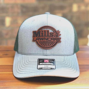 May include: A gray and green trucker hat with the text "Mills Lawncare" in a brown leather patch. The hat has a curved brim and an adjustable snap closure. The Richardson 112 hat is on a wooden surface.