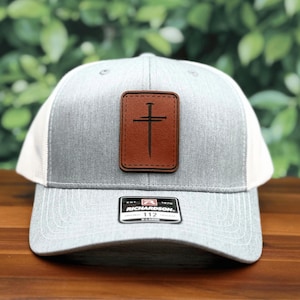 May include: A gray and white trucker hat with a brown leather patch featuring a cross and nail design. The hat has a curved brim and a Richardson 112 tag.