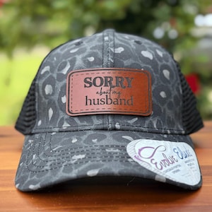 May include: A black and gray trucker hat with a leopard print design. The hat features a brown leather patch with the words "SORRY about my husband" in white lettering. A white sticker with purple text is on the brim.