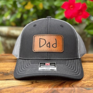 May include: A charcoal gray and light gray trucker hat featuring a rectangular tan leather patch on the front. The patch is engraved with the word Dad in a handwritten style font. A black and white Richardson 112 brand sticker is on the brim.