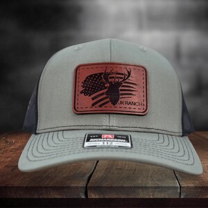 May include: A grey baseball cap with a dark brown rectangular leather patch on the front. The patch features a silhouette of a deer head overlaid on a stylized American flag and the text JK RANCH. A small black and white sticker is on the brim.
