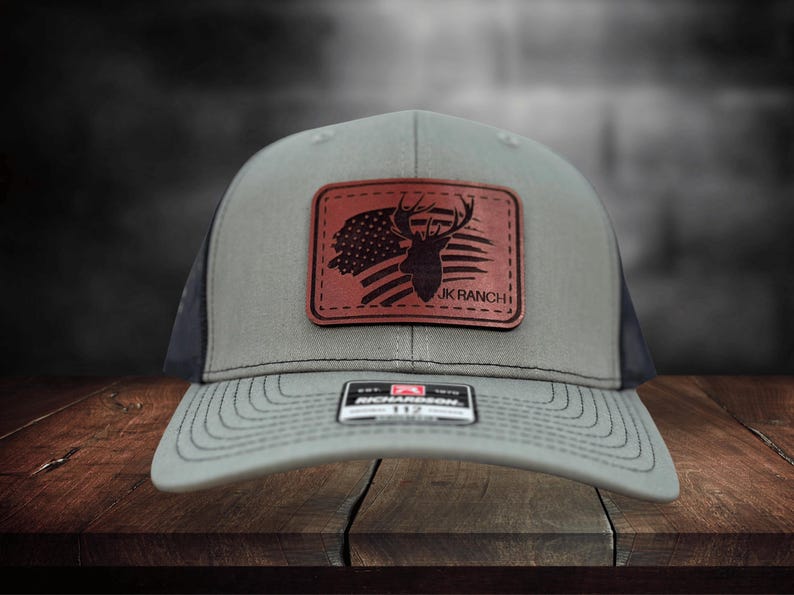 May include: A grey baseball cap with a dark brown rectangular leather patch on the front. The patch features a silhouette of a deer head overlaid on a stylized American flag and the text JK RANCH. A small black and white sticker is on the brim.
