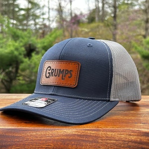 May include: A navy blue baseball cap with a grey mesh back and a rectangular brown leather patch on the front. The patch is embossed with the word GRUMPS in a stylized black font. A small black and white sticker is visible on the curved brim.