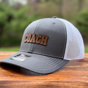 May include: A gray and white trucker hat with a brown leather patch that says "COACH" on it.