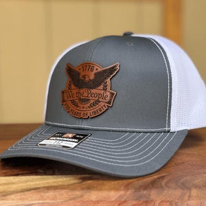May include: A gray and white trucker hat featuring a brown leather patch on the front. The patch displays an eagle graphic, the text 1776, We the People, and 250 YEARS OF LIBERTY. A black Richardson 112 sticker is visible on the curved brim.