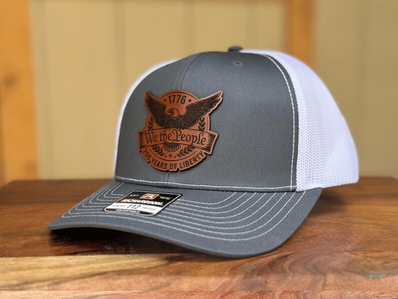 May include: A gray and white trucker hat featuring a brown leather patch on the front. The patch displays an eagle graphic, the text 1776, We the People, and 250 YEARS OF LIBERTY. A black Richardson 112 sticker is visible on the curved brim.
