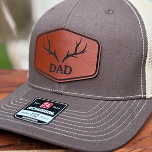 Pu&ograve; includere: Un cappellino trucker grigio antracite e beige con una toppa in pelle marrone sul davanti. La toppa mostra una grafica nera di corna di cervo sopra la scritta DAD. Un adesivo nero sulla visiera riporta la scritta Richardson Original 112 Trucker.